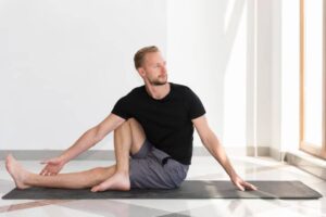 A person in a black t-shirt and gray shorts practices a seated yoga stretch on a mat, surrounded by bright, natural light.