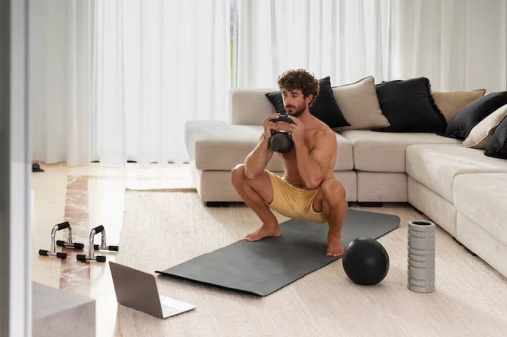 A man performs a squat with a kettlebell on a yoga mat in a modern living room, equipped with fitness equipment and a laptop.