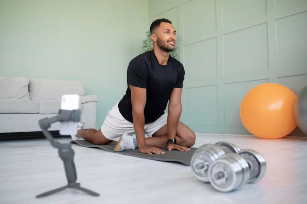 A person practicing yoga on a mat at home, with dumbbells and exercise balls beside them, and a phone on a gimbal for recording.