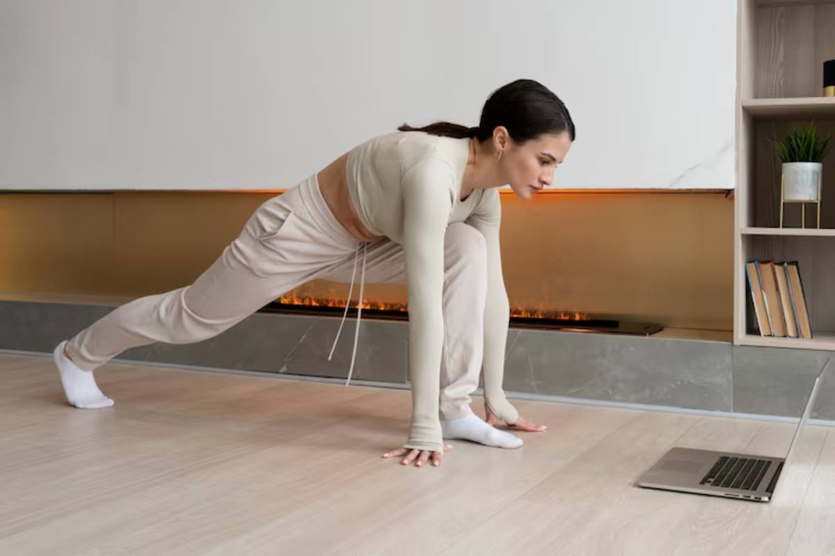 A person in beige activewear performs a lunge exercise at home, with a laptop displaying a workout video in front.