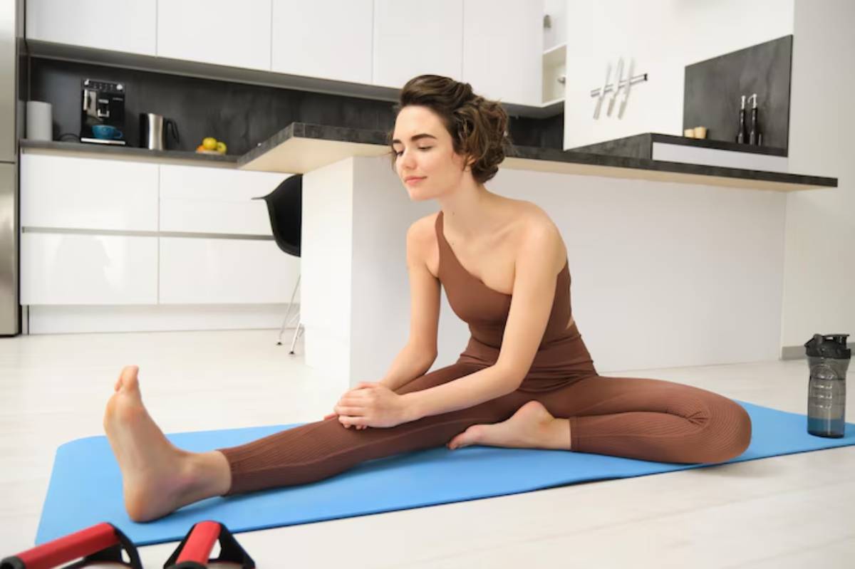 A person in a brown one-shoulder outfit stretches on a blue mat in a modern kitchen, with fitness equipment and a water bottle nearby.