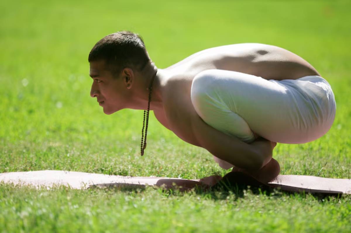 A person practicing yoga in a crouched position on a mat, surrounded by lush green grass under bright sunlight.