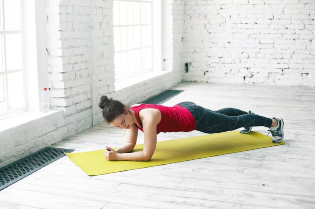 A person performs a plank exercise on a yellow mat in a bright room with white brick walls and large windows.