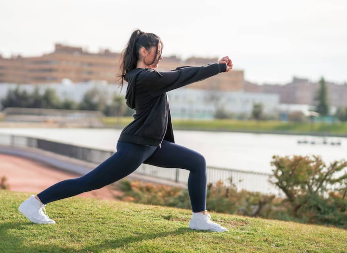 A person performs a lunging exercise on grass with a scenic backdrop, dressed in casual sportswear and white sneakers.