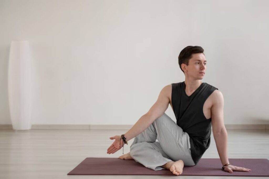 A person in a black sleeveless shirt and light pants sits on a yoga mat, demonstrating a seated twist pose in a calm studio setting.