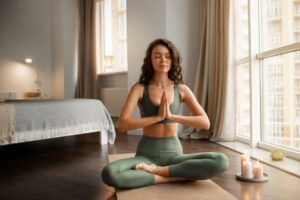 A person in athletic wear sits cross-legged on a mat, practicing yoga in a serene, sunlit room with a bed and candles nearby.