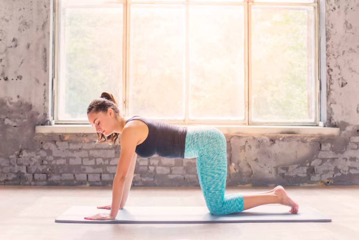  A woman practices yoga on a mat in a sunlit studio with large windows, wearing vibrant teal leggings and a black tank top.