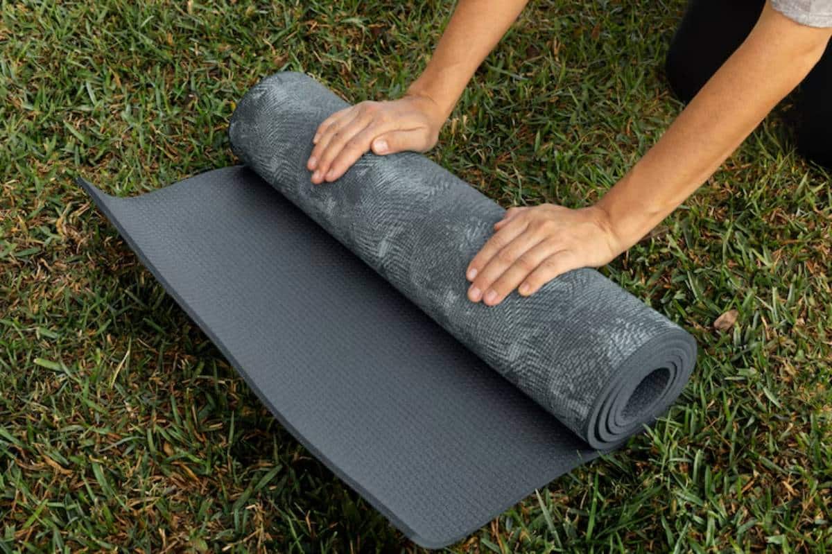 A person rolls out a textured yoga mat on green grass, preparing for a workout or outdoor yoga session.