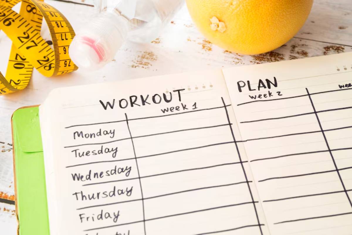 A fitness planner open to workout and plan sections, surrounded by a measuring tape, water bottle, and a grapefruit on a rustic wooden table.
