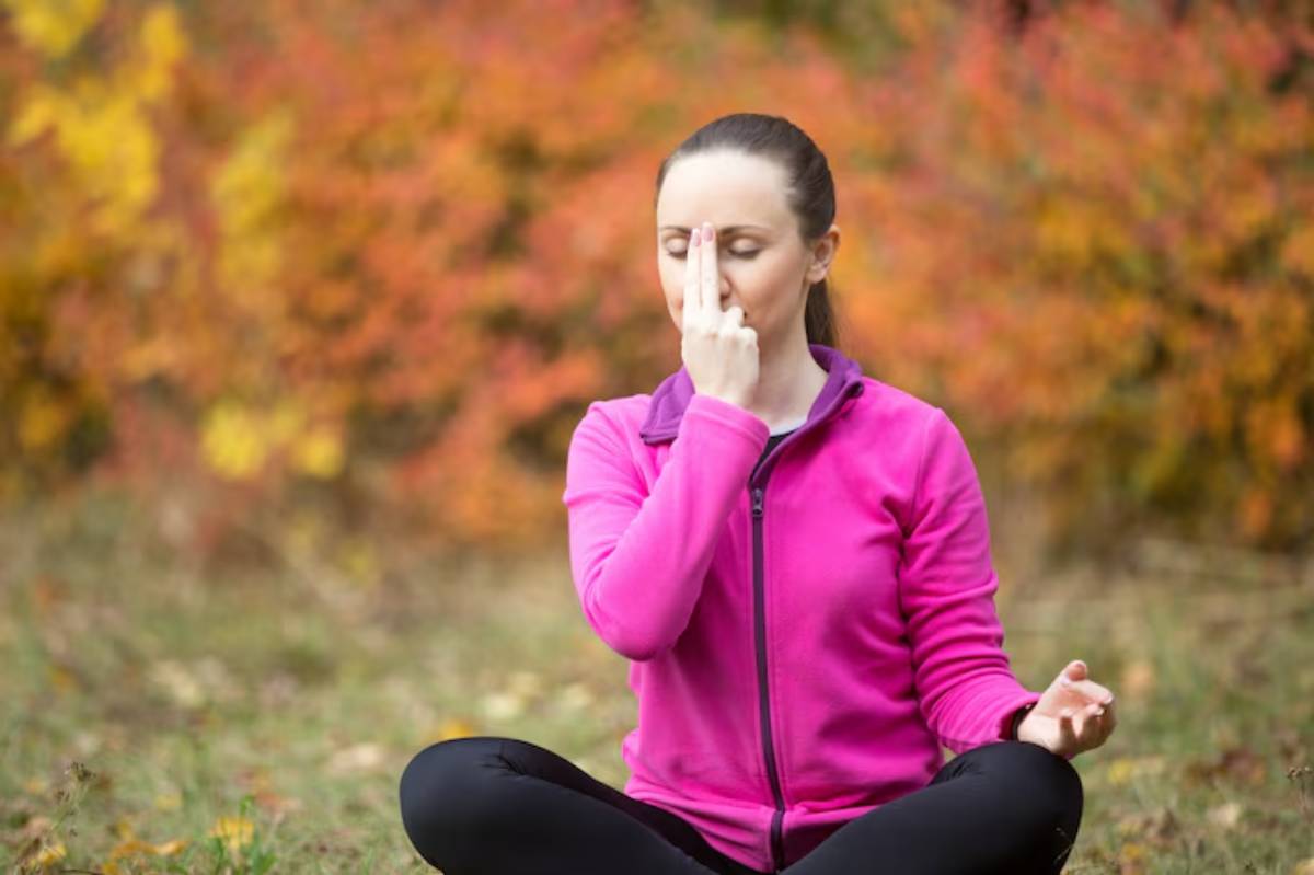 A woman in a pink jacket practices meditation outdoors, surrounded by vibrant autumn foliage in shades of orange and yellow. 