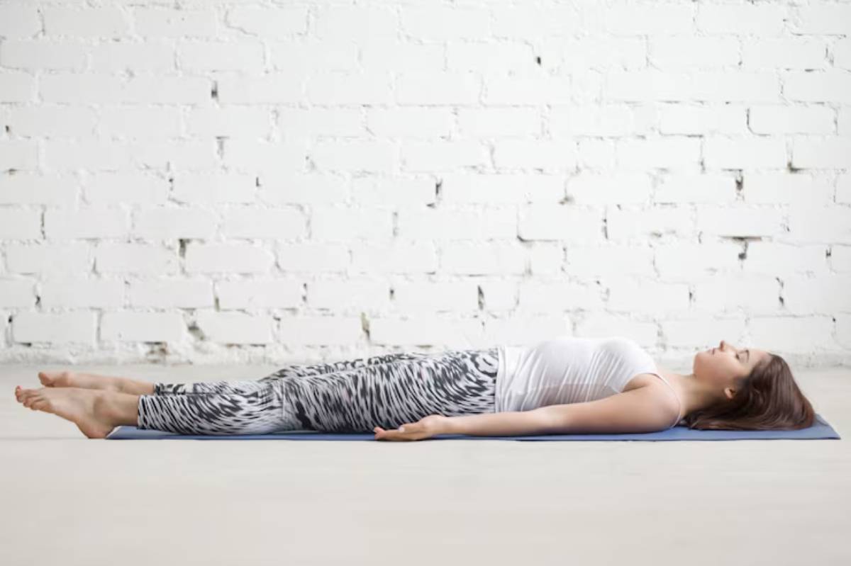  A person lying in Savasana on a yoga mat, wearing a white top and patterned leggings, against a white brick wall.