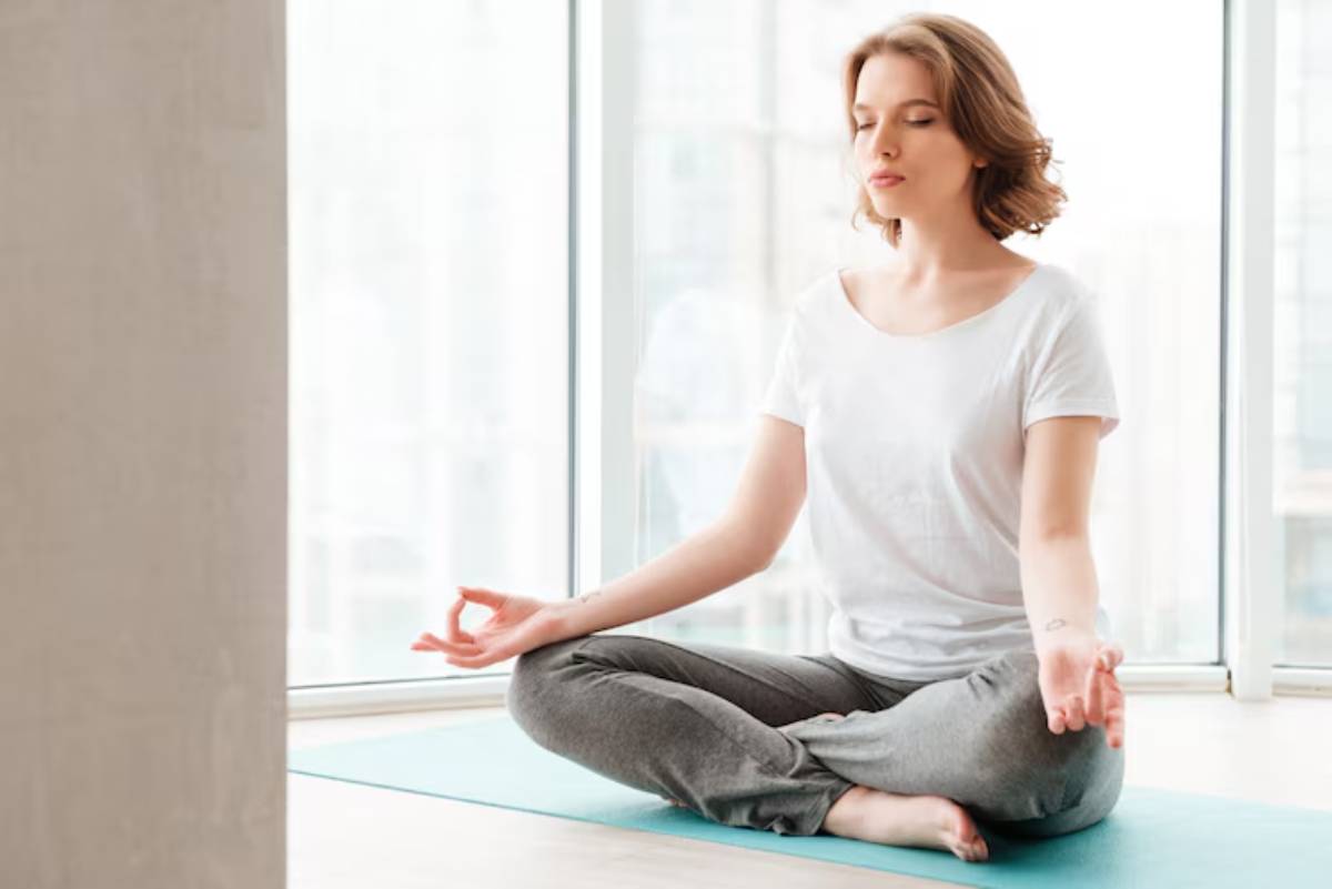 A person sits cross-legged on a yoga mat, practicing meditation in a bright room with large windows.