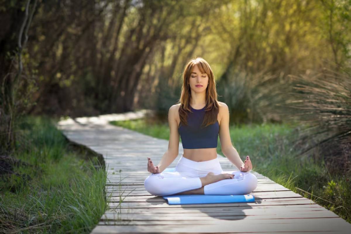 A woman meditates in a serene outdoor setting, seated on a yoga mat along a scenic wooden path surrounded by greenery.