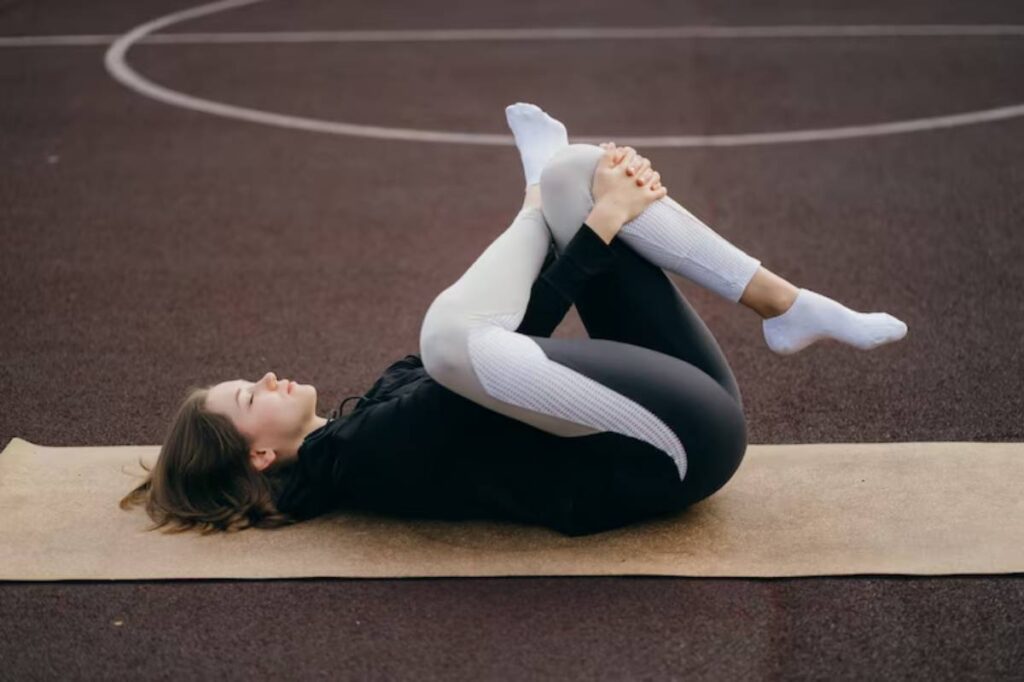 A person in athletic wear performs a yoga pose on a mat on an outdoor court surface. The scene conveys relaxation and focus.