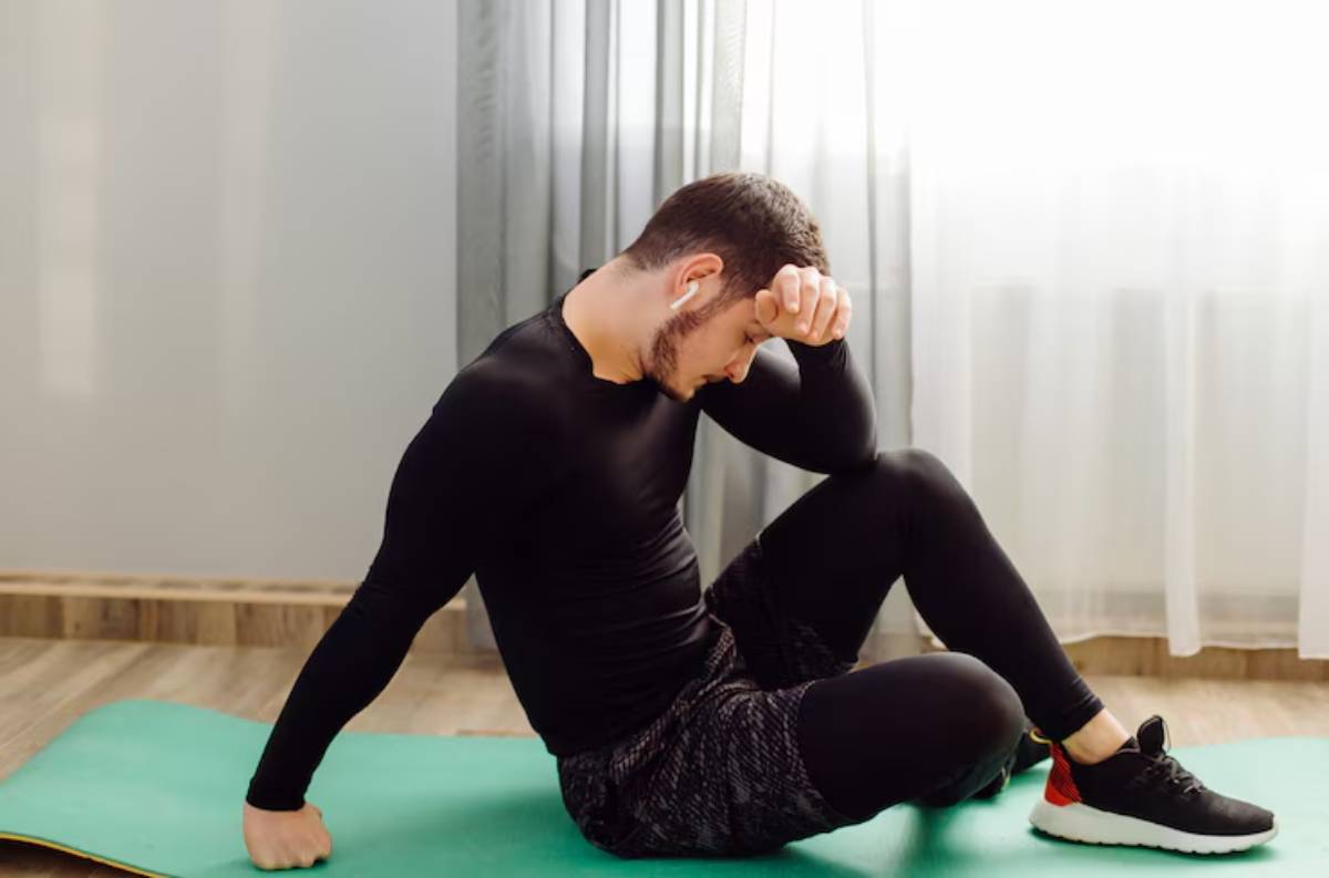 A person in activewear sits on a green yoga mat, resting with a hand on their forehead in a bright, airy room.