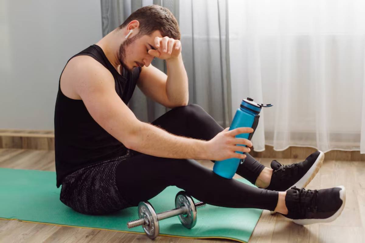  A person in athletic wear sits on a green yoga mat, holding a blue water bottle, with a dumbbell resting beside them.