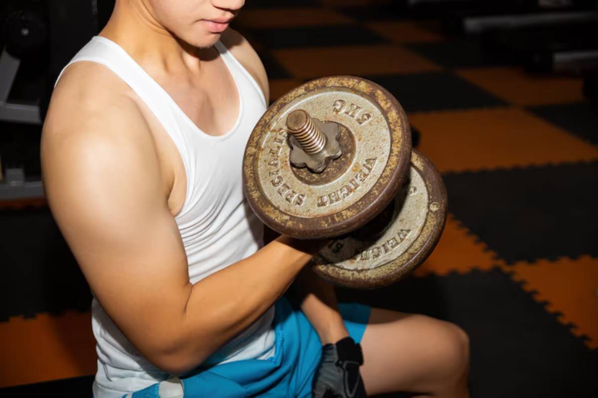  A young man in a tank top lifts a rusty dumbbell in a gym, showcasing his muscular arm against an orange and black floor.