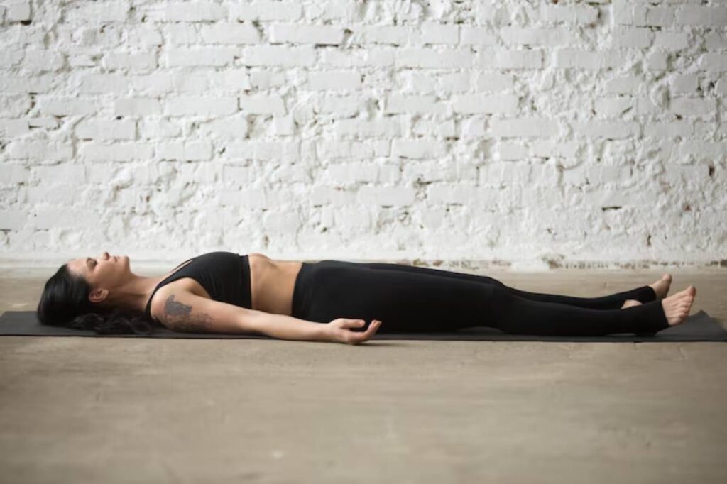 A person lies in a yoga pose on a mat, surrounded by a minimalistic studio with a white brick wall and concrete floor.